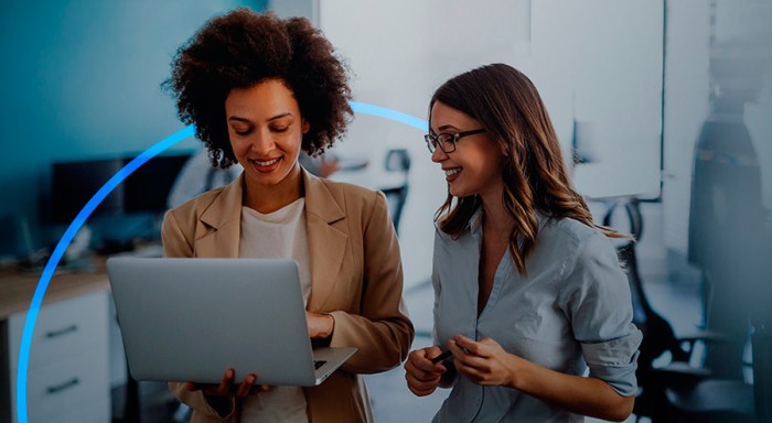 Dos mujeres de pie en una oficina hablan sonrientes mientras miran al ordenador portátil que sostiene la mujer de la izquierda.